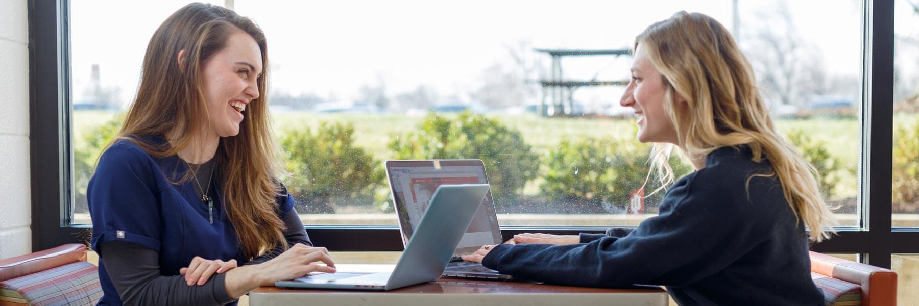 Two female students wear their scrubs and work on their laptops together.
