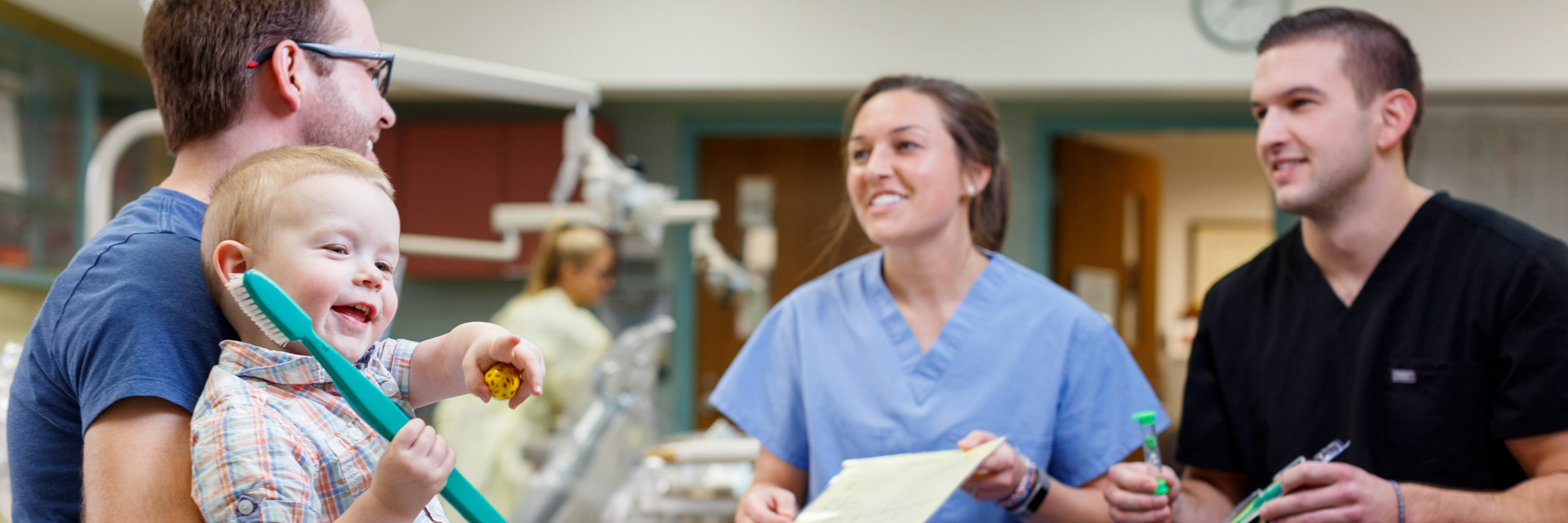 Three dental professionals talk while one holds a small boy playing with a toothbrush. 