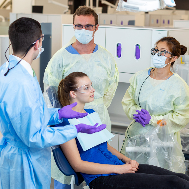 Three dentistry professionals talk as they stand around a young woman sitting in an exam chair.